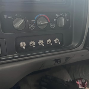 May include: Close-up of a vehicle's dashboard featuring climate controls, including fan speed and air conditioning settings. Below, a panel houses five silver toggle switches, likely for auxiliary functions.