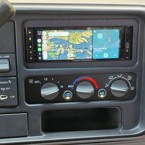 May include: A vehicle dashboard center console featuring a Power Acoustik digital display screen showing a map and music interface, positioned above three circular climate control knobs with labels for fan speed, temperature, and airflow settings.