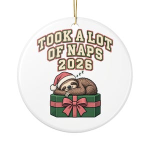 May include: A round white ceramic ornament featuring a sleeping sloth wearing a red Santa hat, resting on a green gift box with a pink ribbon. The text reads TOOK A LOT OF NAPS 2026 in a bold, arched font above the sloth illustration.