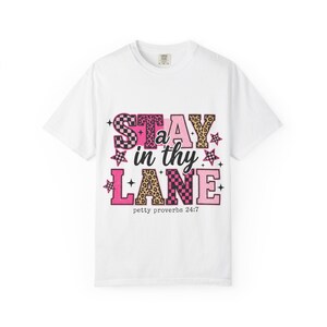 May include: A white short-sleeve t-shirt featuring a graphic design with the text STAY in thy LANE and petty proverbs 24:7. The letters are decorated with pink, black, and brown leopard print and checkered patterns, accented by small star shapes.