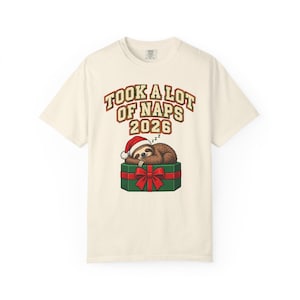 Può includere: Una t-shirt a maniche corte color bianco sporco con la grafica di un bradipo marrone che indossa un cappello da Babbo Natale, mentre dorme su un regalo verde con un nastro rosso. Il testo sopra recita TOOK A LOT OF NAPS 2026 in rosso e verde.