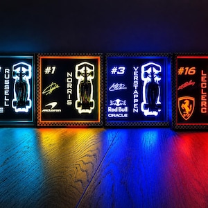 May include: Four illuminated Formula 1 racing art pieces. Each features a driver's number, name, and a car silhouette. The pieces are framed and lit with blue, orange, and red hues. The logos of the teams are also visible.