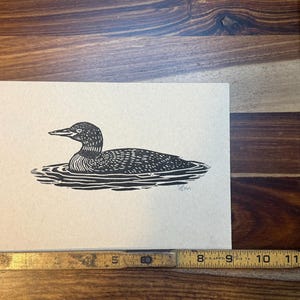 May include: A black and white linocut print of a loon swimming in water. The artwork is on a light tan paper. The print is approximately 8 inches wide and 6 inches tall.