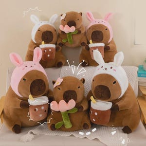 Puede incluir: Un grupo de peluches de capibara marrones, algunos con gorros de conejito rosas o blancos. Algunos sostienen flores rosas o tazas de té con burbujas. Los capibaras tienen ojos marrones oscuros.