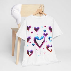 Puede incluir: Camiseta blanca con un diseño de globos en forma de corazón en tonos morados, azules y rojos. Los globos parecen flotar sobre un fondo blanco. Una gorra de béisbol blanca descansa sobre una silla de madera.
