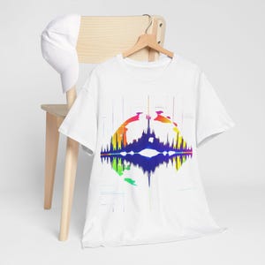 Puede incluir: Camiseta blanca con un diseño gráfico colorido. El diseño presenta una forma abstracta simétrica con tonos vibrantes de amarillo, naranja, verde, azul y morado. La camiseta está colgada en una percha de madera.