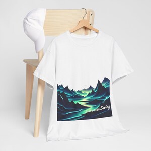 Puede incluir: Camiseta blanca con un gráfico de un paisaje montañoso en tonos turquesa y verde, con el texto "Aurora Swing". Una gorra de béisbol blanca descansa sobre una silla de madera.