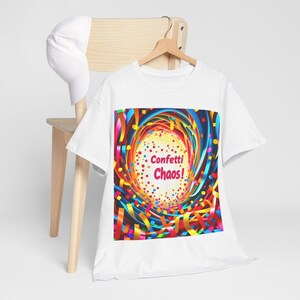 May include: White t-shirt with a colorful graphic design featuring the text "Confetti Chaos!". The design includes swirling ribbons and confetti in various colors, creating a festive and vibrant look. A white baseball cap rests on a wooden chair.