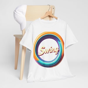 Puede incluir: Camiseta blanca con un diseño circular colorido y la palabra "Swing" en una fuente retro. Una gorra de béisbol blanca descansa sobre una silla de madera detrás de la camiseta, que cuelga de una percha de madera.