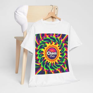 Puede incluir: Camiseta blanca con un diseño abstracto vibrante. El diseño presenta un patrón arremolinado de verde, amarillo, naranja y azul, con el texto "Chaos Play" en un logotipo circular. Una gorra de béisbol blanca descansa sobre una silla de madera.
