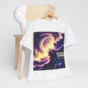 Puede incluir: Camiseta blanca con un diseño cósmico vibrante en tonos amarillos, morados y azules. El gráfico incluye el texto "Galaxy Swing" y "Cosmic Harmony". Una gorra de béisbol blanca descansa sobre una silla de madera.