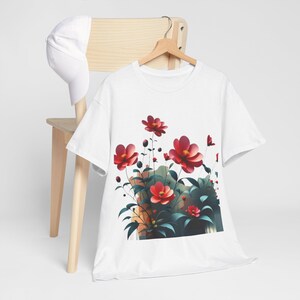 Puede incluir: Camiseta blanca con un diseño floral de flores rojas, hojas verdes y un toque de naranja. La camiseta está colgada en una percha de madera. Una gorra de béisbol blanca descansa sobre una silla de madera.