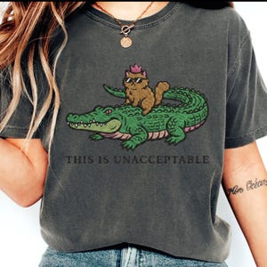 May include: Dark gray t-shirt featuring a cartoon illustration of a cat wearing sunglasses and a crown, riding on a green alligator. The text "THIS IS UNACCEPTABLE" is printed below the illustration.