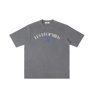 May include: A charcoal gray short-sleeve t-shirt featuring the white arched text LES LÉOPARDS printed across the chest. Below the text is a small square graphic depicting the flag of the Democratic Republic of the Congo with blue, yellow, and red colors.