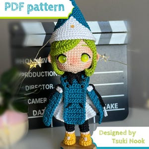 Puede incluir: Una muñeca de ganchillo con cabello verde lima, ojos amarillos grandes y un sombrero puntiagudo azul con borde blanco. La muñeca viste un conjunto azul y blanco con detalles negros y zapatos amarillos sobre una base de madera con la etiqueta Tsuki Nook. Texto: PDF pattern y Designed by Tsuki Nook.