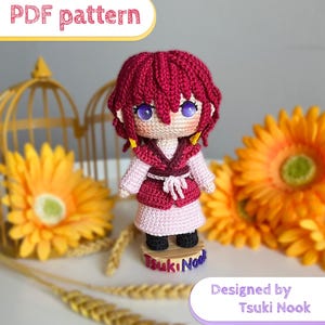 Pode incluir: Boneca de crochê com cabelo vermelho, vestida com uma roupa rosa e bordô. A boneca tem olhos grandes, roxos claros e está em uma pequena base de madeira. O texto "PDF pattern" e "Designed by Tsuki Nook" são visíveis.