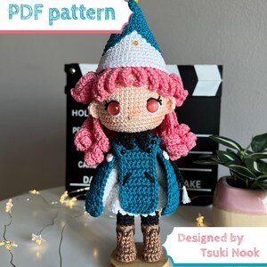 May include: A crochet doll with pink hair, wearing a teal and white pointed hat, a teal cape over a white dress, and brown boots. The doll stands on a wooden base with the text Tsuki Nook. The image includes the text PDF pattern and Designed by Tsuki Nook.