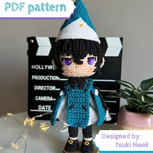 May include: A crochet doll featuring black hair, purple eyes, and a teal and white pointed hat with a yellow tassel. The doll wears a teal tunic over a white garment, black trousers, and black shoes with yellow accents, standing on a wooden base.