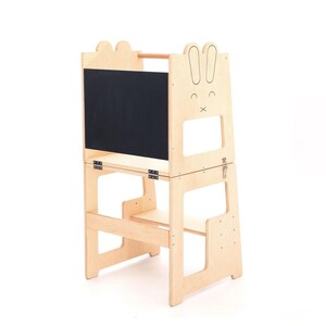 May include: A light-colored wooden learning tower featuring a black chalkboard panel on the front. The side panels are shaped like rabbit ears with engraved facial features. The structure includes a horizontal bar, metal hinges, and a latch.