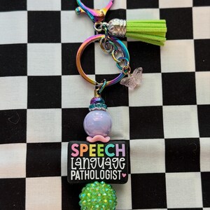 May include: A keyring featuring a rainbow-coloured metal ring and clasp, a lime green tassel, and a small translucent butterfly charm. A black rectangular bead displays the text SPEECH LANGUAGE PATHOLOGIST in colourful letters, flanked by a purple bead and a textured lime green sphere.