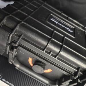 Puede incluir: Maleta negra y rectangular de carcasa dura con una superficie texturizada y un asa. La maleta tiene un cierre de pestillo y un dial con detalles en naranja. El texto "Apex Pro" es visible en la maleta.