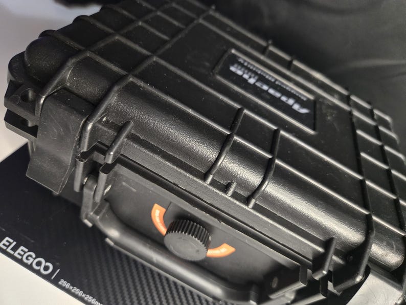 Puede incluir: Maleta negra y rectangular de carcasa dura con una superficie texturizada y un asa. La maleta tiene un cierre de pestillo y un dial con detalles en naranja. El texto "Apex Pro" es visible en la maleta.