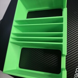 Puede incluir: Objeto rectangular verde brillante impreso en 3D con m&uacute;ltiples divisores horizontales. El objeto tiene forma de caja con la parte superior e inferior abiertas. Se coloca sobre una superficie negra con un patr&oacute;n de fibra de carbono.
