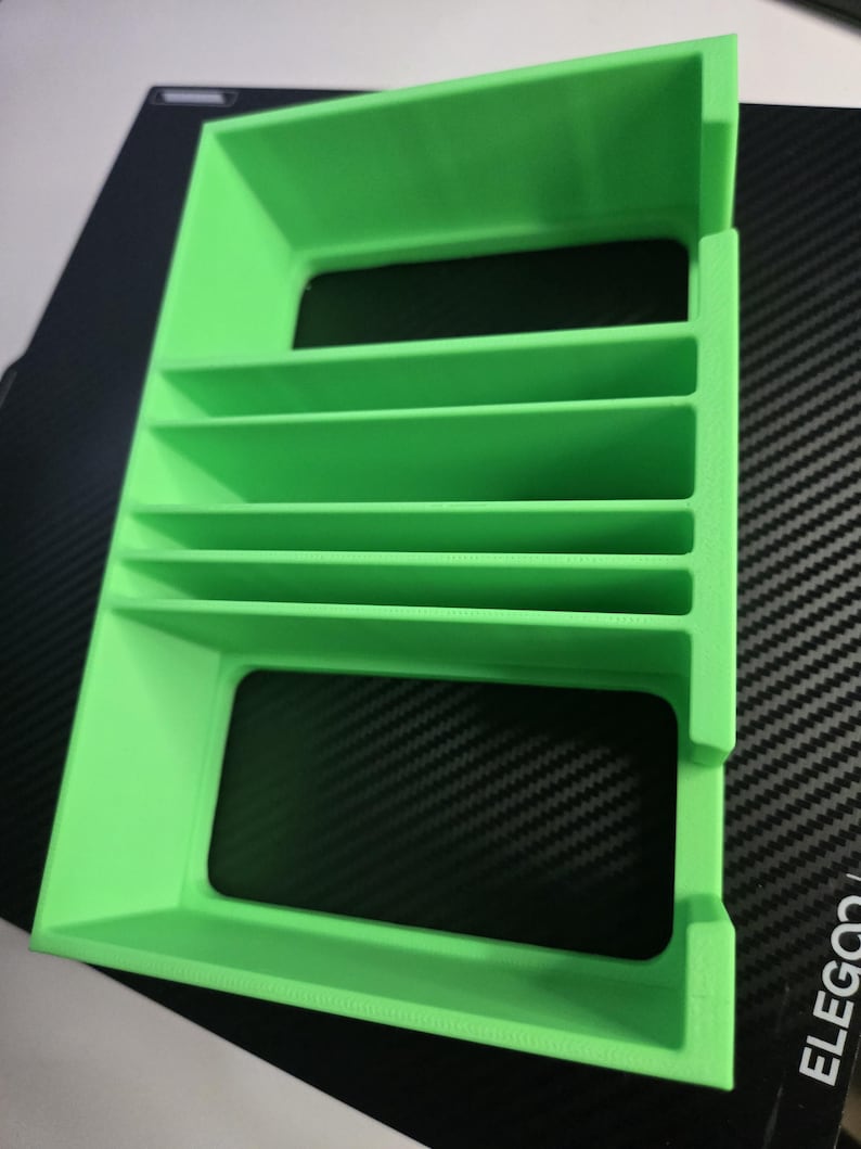 Puede incluir: Objeto rectangular verde brillante impreso en 3D con m&uacute;ltiples divisores horizontales. El objeto tiene forma de caja con la parte superior e inferior abiertas. Se coloca sobre una superficie negra con un patr&oacute;n de fibra de carbono.
