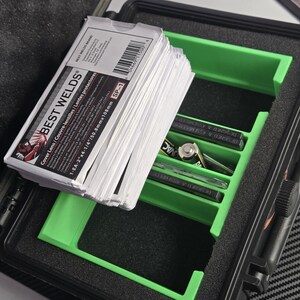 Puede incluir: Un malet&iacute;n negro abierto con un inserto verde que contiene pilas y otros art&iacute;culos. Una pila de tarjetas blancas con el texto "BEST WELDS" es visible. El malet&iacute;n est&aacute; sobre una superficie estampada.