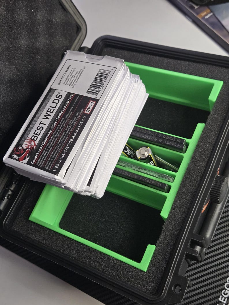 Puede incluir: Un malet&iacute;n negro abierto con un inserto verde que contiene pilas y otros art&iacute;culos. Una pila de tarjetas blancas con el texto "BEST WELDS" es visible. El malet&iacute;n est&aacute; sobre una superficie estampada.