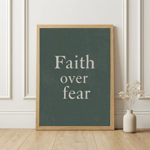 Peut inclure: Une affiche encadrée avec les mots "Faith over fear" en lettres blanches sur un fond vert foncé. Le cadre est en bois brun clair. Un vase blanc avec des fleurs séchées est à droite du cadre sur un sol en bois.