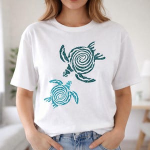 May include: White t-shirt featuring two stylized sea turtles in teal glitter. The turtles have a spiral design on their shells and are positioned one above the other. The shirt is a casual, everyday wear item.