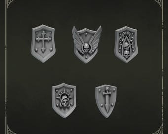 May include: A set of five grey miniature shoulder shields labeled Shoulder shields 2. The shields feature raised designs including a cross, a winged skull, two stacked skulls with scrollwork, a hooded skull with a halo, and a sword with rivets.