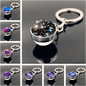 May include: A collection of keychains featuring a celestial design. Each keychain has a round, glass-like pendant with a galaxy or starry night pattern in shades of blue, purple, and black, set in a silver-colored metal frame. The keychains are attached to a silver key ring.