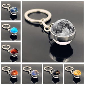 May include: A collection of planet-themed keychains. Each keychain features a spherical charm depicting a planet, including Earth, the Moon, and others. The charms are encased in a silver-colored metal setting and attached to a keyring.
