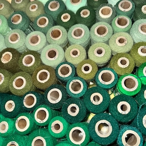 May include: A collection of spools of thread in various shades of green, from light pastel to deep forest. The spools are arranged closely together, creating a gradient effect. The wooden cores of the spools are visible.