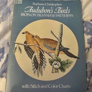 May include: A vintage book titled "Audubon's Birds" by Barbara Christopher. The cover features a detailed illustration of two birds perched on a branch within a circular frame. The book includes iron-on transfer patterns and stitch and color charts.