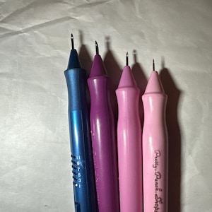 Puede incluir: Cuatro herramientas de aguja de perforación en tonos de azul, morado y rosa. La herramienta azul tiene marcas de medición. La herramienta rosa tiene el texto "Pretty Punch Shoppetters" impreso. Estas herramientas se utilizan para bordar con aguja de perforación.