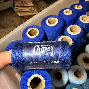 May include: A collection of blue spools of Cameo needlepunch thread. The spools are various shades of blue, with a central cardboard core. The label on the spool reads "Cameo Products by Inc. Orlando, FL 32859".