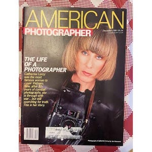 May include: The December 1988 issue of American Photographer magazine features a portrait of Catherine Leroy on the cover. She has blonde hair and wears a black leather jacket with a camera strap. The text reads The Life of a Photographer.