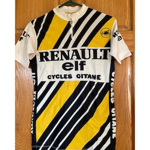 May include: A short-sleeve cycling jersey featuring a yellow, black, and white diagonal striped pattern. The front displays the text RENAULT elf CYCLES GITANE in black lettering, a small scorpion logo, and a white collar with a partial front zipper.