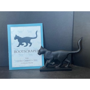 May include: A black cast iron boot scraper in the shape of a cat, with a matching card that reads "BOOTSCRAPER" and "THE CURIO CABINET CATS COLLECTION". The cat is in a walking pose.