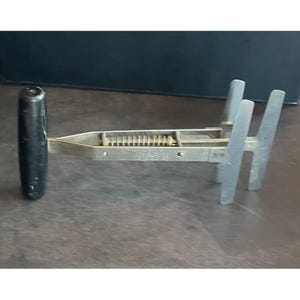 May include: A metal tool featuring a black cylindrical handle attached to a horizontal frame with a central spring mechanism and two vertical prongs at the opposite end. The device has a silver-toned finish and a mechanical structure.