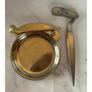 Könnte beinhalten: Ein Vintage-Golf-Aschenbecher-Set. Das Set enthält einen runden, goldfarbenen Aschenbecher mit Golfschläger- und Ball-Detail sowie ein passendes Divot-Werkzeug. Der Aschenbecher hat einen Durchmesser von etwa 10 cm.