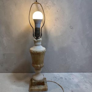 May include: A table lamp featuring a carved alabaster stone base with a classic urn-shaped body, a square pedestal, and a metal harp fixture holding a single illuminated white LED light bulb with a visible power cord extending from the base.