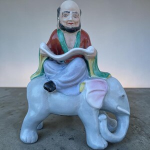 Peut inclure: Une figurine en céramique représentant un personnage chauve et barbu avec un bouc foncé, vêtu d'une robe rouge et verte, assis en tailleur sur un éléphant bleu clair. Le personnage tient un parchemin blanc, et l'éléphant a des oreilles roses.