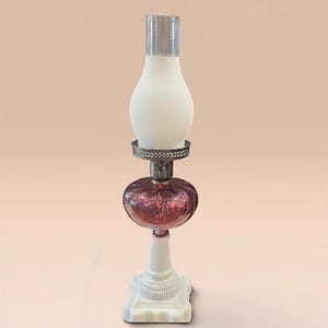 Könnte beinhalten: Eine Tischlampe mit einem quadratischen Fuß aus weißem Milchglas, einem bauchigen Reservoir aus dunkelviolettem Glas und einem Schirm aus mattiertem weißem Glas. Ein silberfarbener Metallring mit geprägtem Muster verbindet die einzelnen Teile.
