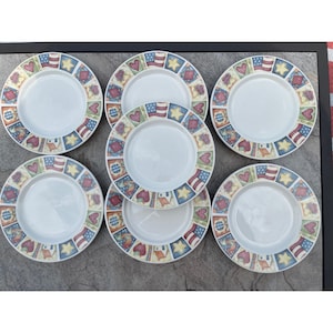 May include: A set of seven white ceramic plates with a decorative border featuring repeating patterns of red hearts, yellow stars, blue and white checkered squares, American flags, and small houses in a grid arrangement around the rim.