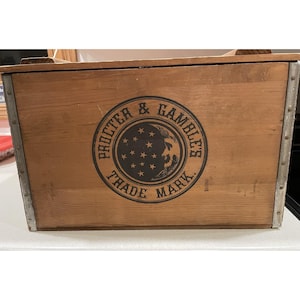 Puede incluir: Caja de madera con esquinas reforzadas con metal. La parte frontal de la caja presenta un emblema circular con las palabras "PROCTER & GAMBLE'S TRADE MARK" que rodea un diseño de una cara y estrellas.