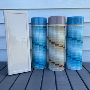 May include: A rectangular off-white plastic case stands next to three cylindrical Thermos brand vacuum-insulated bottles. Two bottles feature a blue grid pattern, while the center bottle displays a brown and beige grid pattern with matching lids.